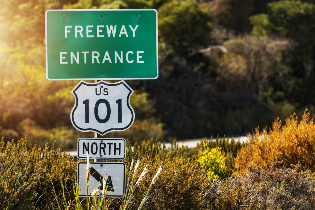 U.S. Route 101 highway sign at a scenic outdoor location, with a green roadside sign indicating "Freeway Entrance" amidst natural foliage and sunlight.