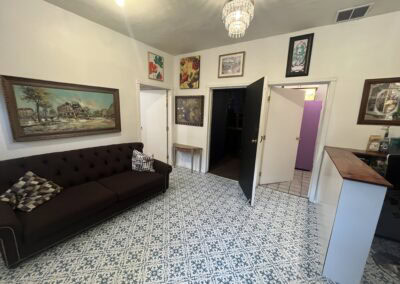 Photo of an art-filled living room with a dark brown tufted sofa, framed paintings and colorful wall art, a chandelier, patterned tile flooring, and a doorway leading to another room.