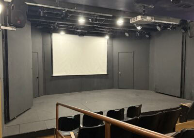Empty black box theater stage with a large white projection screen and professional audio and lighting equipment, perfect for theatrical productions, performances, and stagecraft events.