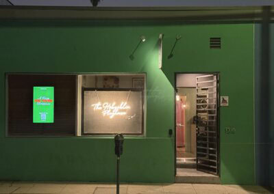 Modern green storefront of The StageCrafts theater, featuring illuminated neon sign and digital marquee promoting holiday shows, with secure entrance and urban street setting.